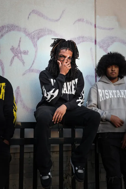 Man with locs and glasses in a black Risktaker hoodie, sitting on a railing in front of a graffiti wall