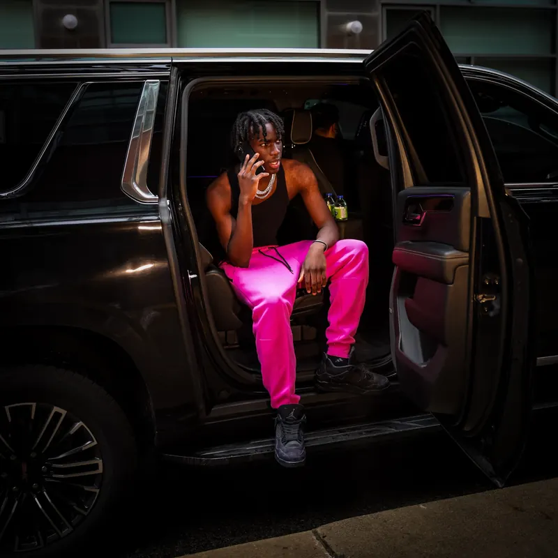 Man in hot-pink sweatpants on a phone call, sitting in the open rear door of a black SUV