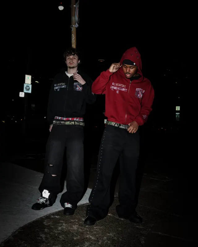 Two men at night in matching black and red “All Weapons Formed Against Me” zip hoodies
