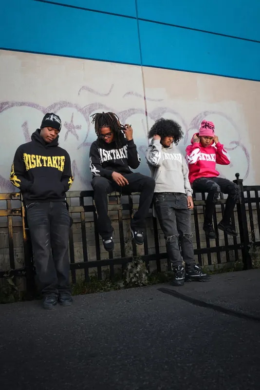 Four people in Risktaker hoodies on a black railing in front of a graffiti wall